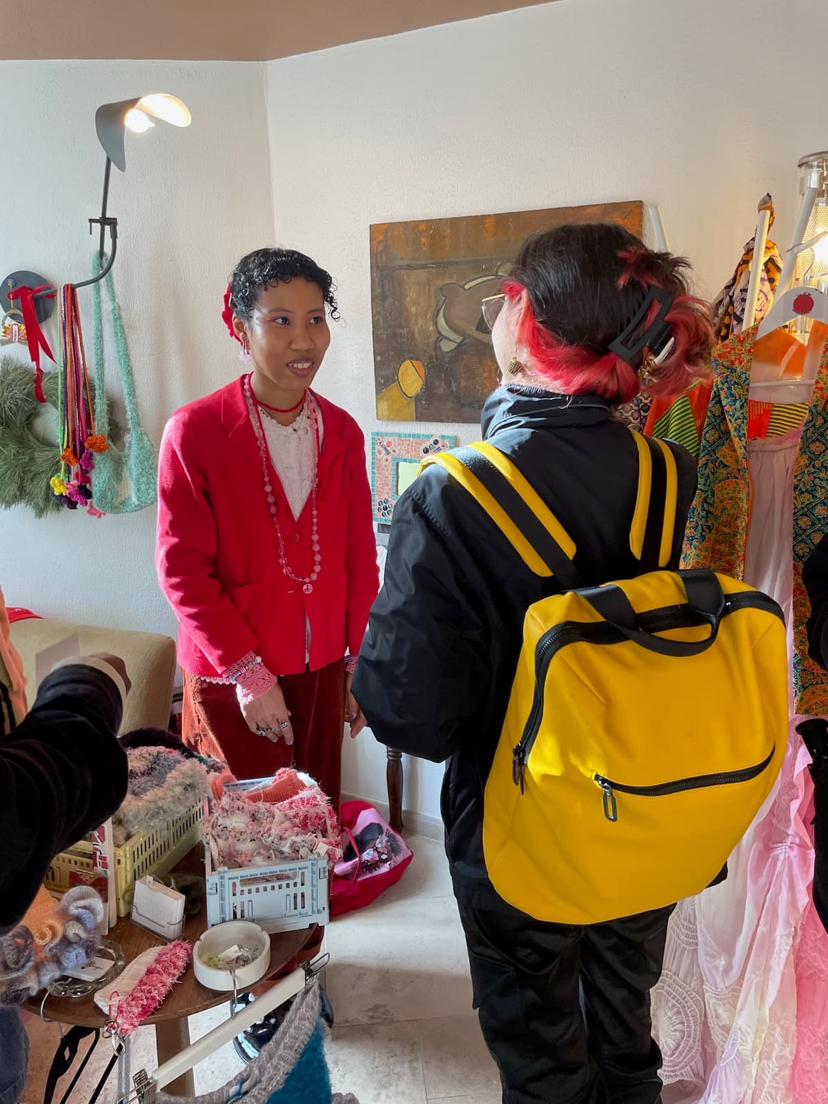 A maker in a red blazer talking with a visitor beside a rail of clothes